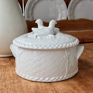 以下が含まれることがあります： 白いセラミック製の蓋付きサービングディッシュ。編みかごのデザインと、蓋に2羽の鳥の置物があります。このディッシュには取っ手があり、他の白いセラミック製の食器とセットになっています。