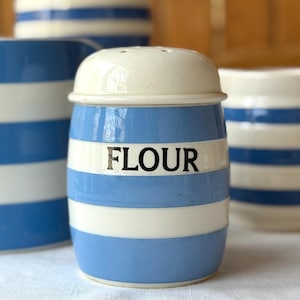 Peut inclure: Un pot à farine en céramique à rayures bleues et blanches, avec un couvercle blanc. Le mot "FLOUR" est imprimé en noir sur le devant. D'autres ustensiles de cuisine assortis sont visibles en arrière-plan.