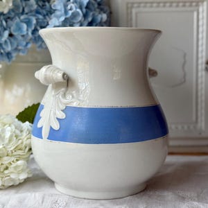 May include: A white ceramic vase with a blue horizontal stripe and decorative handles. The vase features a leaf design and a bulbous base. The vase is a decorative item for the home.