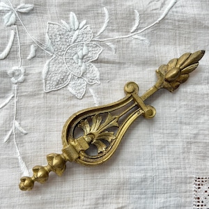 Peut inclure: Élément décoratif en laiton antique orné de motifs floraux et de feuilles. La pièce a une forme symétrique avec des gravures détaillées et une teinte dorée. Il repose sur un tissu blanc brodé.