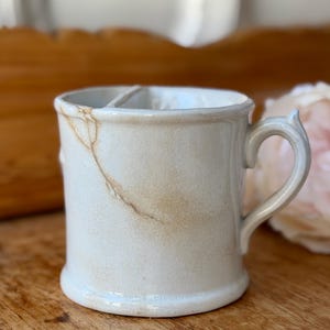 Op de afbeelding: Een crèmekleurige keramische scheerkop met een handvat en twee interne compartimenten. De mok heeft een scheur langs de zijkant en een decoratief handvat. De mok staat op een houten oppervlak.