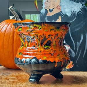 Puede incluir: Un caldero de cerámica negro y naranja con tres patas. El caldero tiene un efecto de esmalte goteado en naranja, amarillo y negro. Una calabaza naranja está en el fondo, junto con una decoración con temática de brujas.