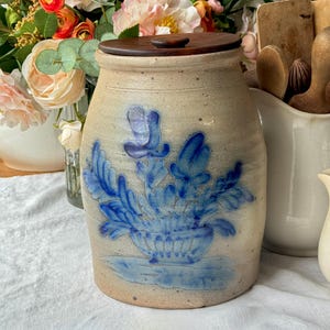 May include: A beige ceramic jar with a wooden lid. The jar features a blue floral design. The jar is cylindrical with a slightly wider base. The lid is round and dark brown. The jar is likely used for kitchen storage.