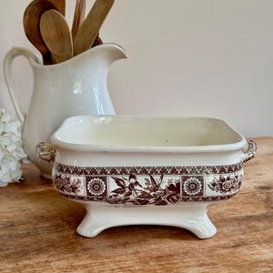 Puede incluir: Fuente rectangular de cerámica color crema con un estampado floral y de pájaros marrón. La fuente tiene una base con patas y asas pequeñas. Una jarra blanca con cucharas de madera está en el fondo.