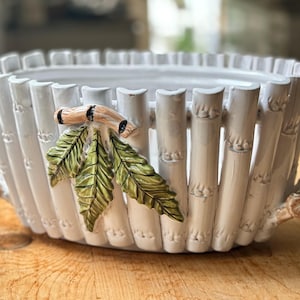 May include: White ceramic planter with a bamboo design. The planter features vertical bamboo-like segments and is adorned with a bamboo branch and green leaves. The planter is designed to hold plants and flowers.