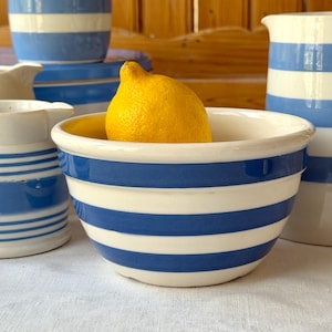 May include: A white ceramic bowl with blue horizontal stripes, holding a bright yellow lemon. Other matching ceramic pieces, including pitchers and bowls, are visible in the background. The items are arranged on a white surface.