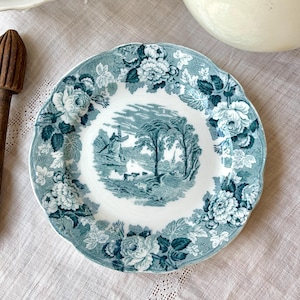 May include: A round, antique-style plate with a scalloped edge. The plate features a teal and white floral border and a central landscape scene with a windmill, trees, and a church. The plate is made of ceramic.