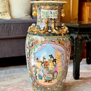 May include: A large, ornate ceramic vase with a wide mouth and a bulbous body. The vase features a detailed, hand-painted scene of figures in a garden, with gold accents and floral patterns. The vase is approximately 18 inches tall.