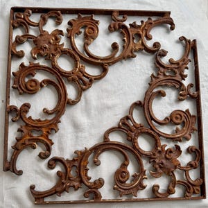May include: Ornate, rusty brown metal corner bracket with scrolling floral and circular designs. The bracket is square with a missing corner, showcasing detailed craftsmanship. Ideal for decorative or restoration projects.
