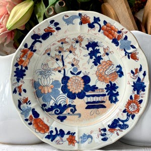May include: A round ceramic plate with a scalloped edge, featuring a white base decorated with intricate floral and botanical patterns in shades of deep blue and vibrant orange, including depictions of stylized flowers and foliage.