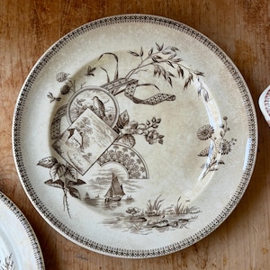 May include: A cream-colored ceramic plate with a brown transferware design. The plate features a detailed landscape scene with a sailboat, bird, and floral elements. The rim has a decorative border.