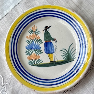 May include: A round, hand-painted ceramic plate with a yellow rim and blue stripes. The plate features a central illustration of a person in traditional attire, holding a rope, with floral and plant motifs. The plate is on a white surface.