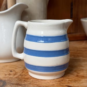 May include: A ceramic creamer pitcher featuring a cream-colored base with three horizontal blue stripes. The pitcher has a rounded body, a small pouring spout, and a single side handle. The surface has a smooth, glossy finish throughout.