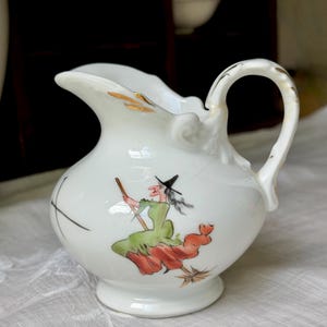 May include: A white porcelain creamer pitcher featuring a hand-painted illustration of a witch in a green dress and black hat flying on a broomstick. The pitcher has a curved handle, a scalloped rim, and gold-painted accents on the handle and rim.