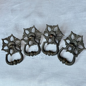 Puede incluir: Cuatro tiradores de cajón antiguos de bronce con diseño floral. Cada tirador presenta una placa trasera en forma de flor y un asa curvada. Los tiradores están dispuestos sobre una superficie blanca, mostrando sus intrincados detalles y pátina envejecida.