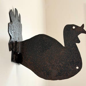 May include: A rustic, metal turkey silhouette wall decor. The dark brown, weathered finish gives it an antique look. The turkey is mounted on a white wall, showcasing its unique shape and design. A decorative piece for a farmhouse or country-themed home.
