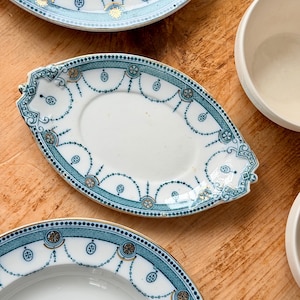 May include: Overhead shot of antique white porcelain plates with a teal and gold decorative border. The plates are oval and round, with a detailed design of connected dots and floral accents. The plates are on a wooden surface.