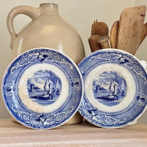 Puede incluir: Dos platos de porcelana azul y blanco con un borde decorativo y una imagen central de una mujer en un jardín.