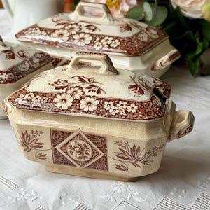 May include: Cream-colored ceramic serving dishes with brown floral and leaf patterns. The lids feature handles and floral designs. The dishes have a rectangular shape with handles on the sides. The set includes a large serving dish and two smaller dishes.