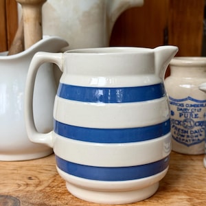 May include: Cream-colored ceramic pitcher with horizontal blue stripes. The pitcher has a handle and a spout for pouring. Other ceramic items are in the background, including a mortar and pestle and a small jar with text.