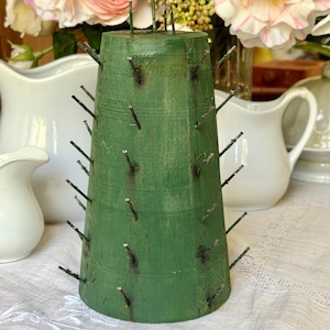 May include: A conical wooden display stand painted in a matte green finish, featuring numerous thin metal spikes protruding from the surface in a spiral pattern, designed for holding small items or ornaments in an upright position.