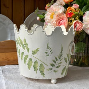May include: A white metal planter with a scalloped top edge and three small ball feet. The exterior features a green fern leaf print and two side ring handles. The container is positioned in front of a bouquet of pink and orange flowers.
