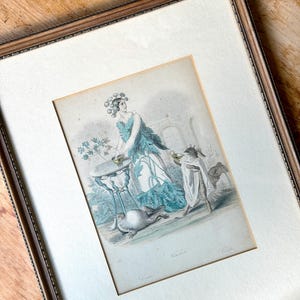 May include: A framed vintage-style print featuring a figure in a teal leaf-patterned garment standing at a small table, accompanied by a small animal wearing a white cloak and a floral headpiece, with a larger animal resting on the ground nearby.