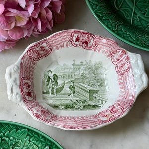 May include: A ceramic serving dish with a scalloped edge and two side handles. The rim features a red decorative pattern, while the center displays a green monochromatic landscape illustration of figures near a stone structure and a body of water.