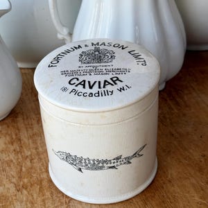 Puede incluir: Tarro de caviar de cerámica color crema con una ilustración de esturión negro. La tapa presenta el texto "FORTNUM & MASON LIMITS" y "CAVIAR 181 Piccadilly W.I." en negro. Jarrones de cerámica blanca en el fondo.