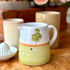 May include: A white ceramic pitcher with a handle, featuring a hand-painted design of a tree and sheep on a green and blue background. Other ceramic kitchenware is in the background, including a juicer and two beige mugs. The pitcher is on a wooden surface.