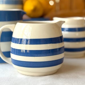 Puede incluir: Jarra de cerámica color crema con rayas horizontales azules. La jarra tiene un asa curva y un pico vertedor. Otras jarras con diseños similares están en el fondo. Las jarras son probablemente para servir bebidas.