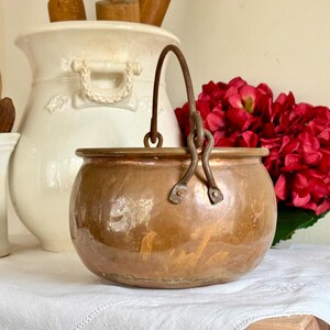 May include: A vintage copper pot with a rustic iron handle. The pot has a rounded shape and a slightly tarnished finish.