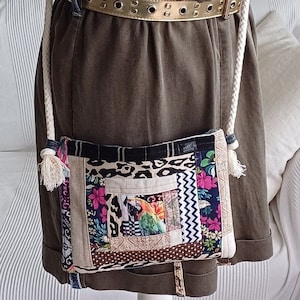 Könnte beinhalten: Eine Patchwork-Tasche aus Stoff mit einem weißen Kordelriemen und einer goldenen Gürtelschnalle. Die Tasche hat verschiedene bunte Muster, darunter Blumen, Tiermuster und geometrische Designs.