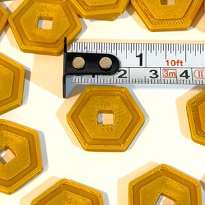 May include: Several gold-colored, hexagonal tokens with a square center hole are arranged on a white surface. A measuring tape shows the tokens are approximately 1 inch in diameter. The tokens have a layered design.