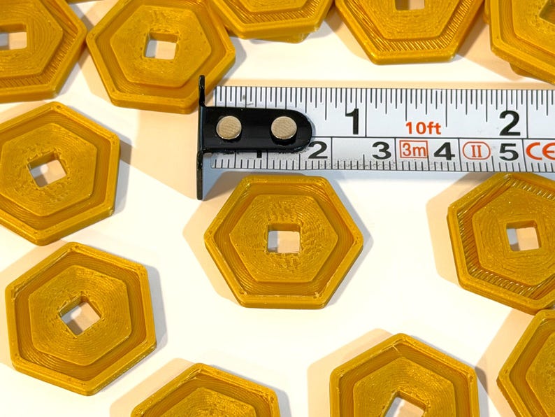 May include: Several gold-colored, hexagonal tokens with a square center hole are arranged on a white surface. A measuring tape shows the tokens are approximately 1 inch in diameter. The tokens have a layered design.
