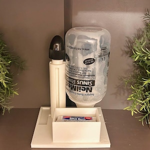 Könnte beinhalten: Ein weißes Nasenspülsystem aus Kunststoff mit einer transparenten Flasche und schwarzem Verschluss. Die Flasche ist mit "NeilMed Sinus Rinse" beschriftet. Eine kleine Box enthält Päckchen. Zwei Topfpflanzen mit grünem Laub flankieren das System.