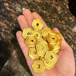 May include: A handful of gold-colored, hexagonal coins with a square cut-out in the center. The coins have a textured surface and are stacked together. The coins are likely for a board game or collectible.