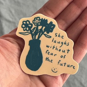 May include: A beige sticker with a hand-drawn illustration of a vase with flowers. The text "she laughs without fear of the future" is written in black cursive. A smiley face is drawn below the text.