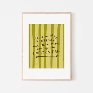 May include: A framed art print with a light wood frame. The artwork features a yellow-green background with vertical stripes. Black handwritten text reads "Focus on the VERTICAL and He'll take care of the HORIZONTAL."