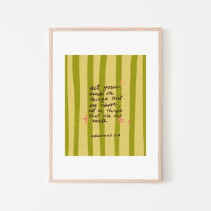 May include: A framed art print with a light wood frame. The artwork features a yellow and green striped background with handwritten text that reads "set your minds on things that are above, not on things that are on earth." Pink star accents are also included.