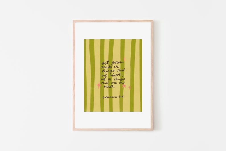 May include: A framed art print with a light wood frame. The artwork features a yellow and green striped background with handwritten text that reads "set your minds on things that are above, not on things that are on earth." Pink star accents are also included.