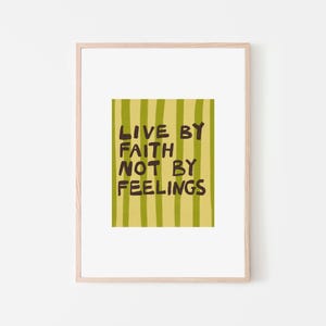 May include: A framed art print with the text "LIVE BY FAITH NOT BY FEELINGS." The artwork features a yellow-green striped background with the text in dark brown. The frame is a light wood color, and the background is white.