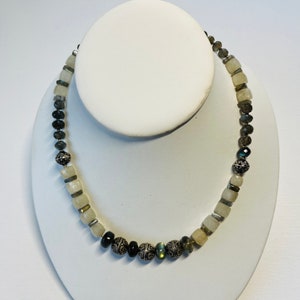 May include: A beaded necklace with a variety of stone beads, including light yellow, dark gray, and silver-toned beads. The necklace is displayed on a white jewelry stand. The beads are various shapes and sizes.