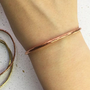 May include: A hammered copper bracelet is worn on a wrist. The bracelet has a textured surface and a simple, elegant design. Other similar bracelets in gold and copper are visible in the background.