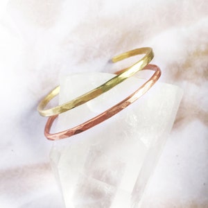 May include: Two hammered metal cuff bracelets. One is gold-toned, the other is copper-toned. The bracelets are stacked on top of each other, with a textured surface. The background is a soft, neutral colour.