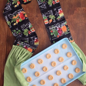 May include: A blue baking tray with baking parchment and a selection of round biscuits. The baking tray is on a wooden surface. A green tea towel with a black and white fabric strip featuring a garden theme and the text "Garden to Table" is on the left of the image.