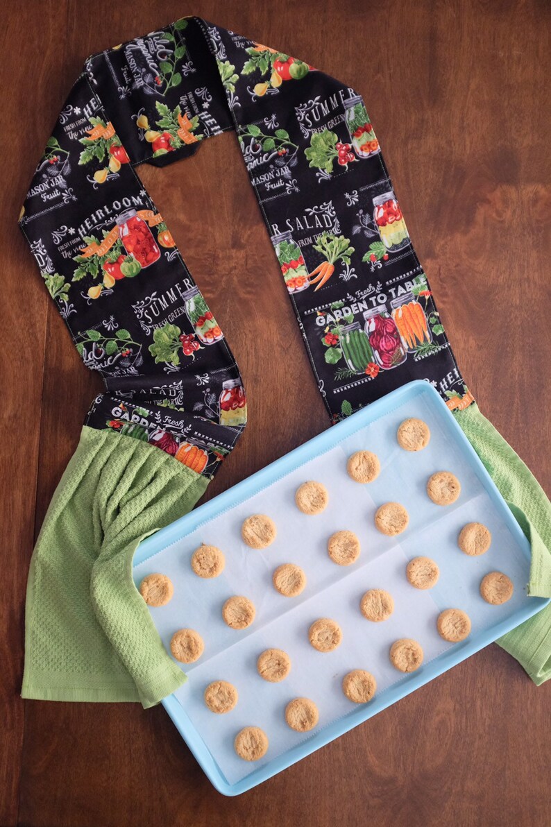 May include: A blue baking tray with baking parchment and a selection of round biscuits. The baking tray is on a wooden surface. A green tea towel with a black and white fabric strip featuring a garden theme and the text "Garden to Table" is on the left of the image.