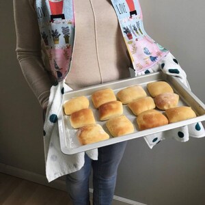 May include: A baking tray filled with freshly baked rolls, held by a person wearing a floral print tea towel and a beige jumper. The towel has the words "I LOVE" printed on it.