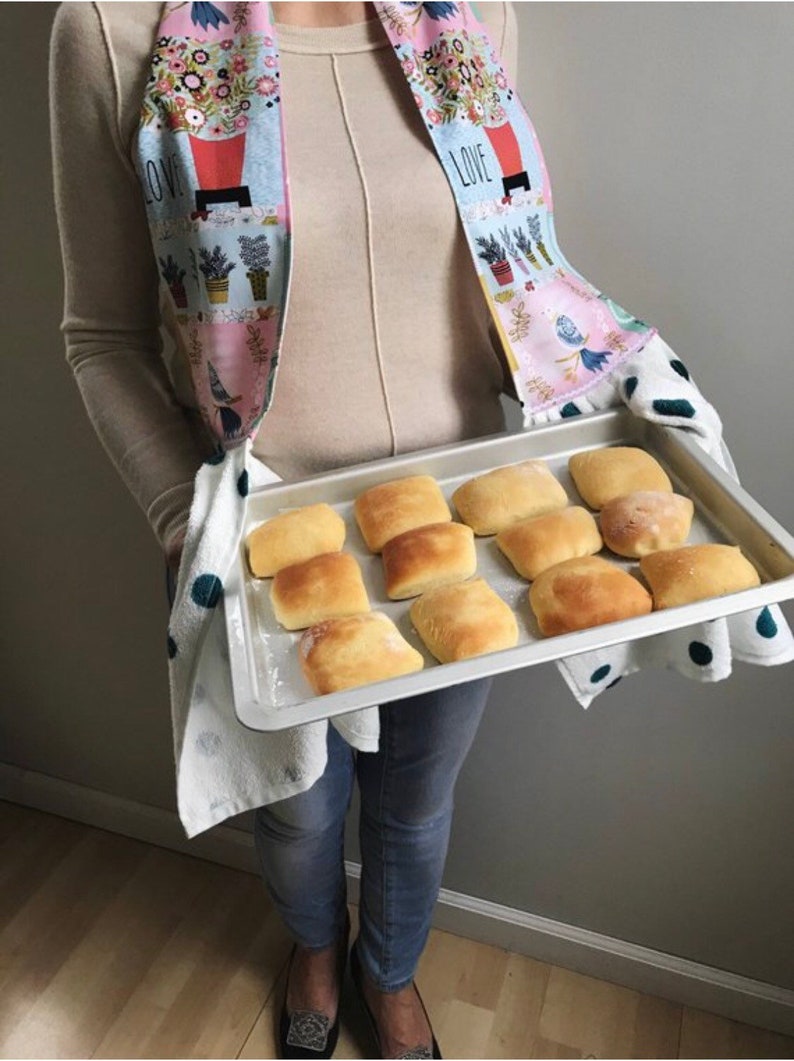 May include: A baking tray filled with freshly baked rolls, held by a person wearing a floral print tea towel and a beige jumper. The towel has the words "I LOVE" printed on it.
