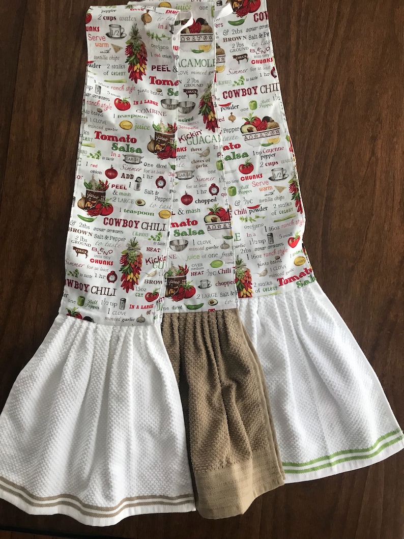May include: Two white kitchen towels with brown and green trim. The towels are decorated with a colourful print of various ingredients and instructions for making salsa and cowboy chili. The text includes "Tomato Salsa", "Cowboy Chili", "Kickin' Guacamole", and "One diced onion".